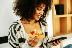 Mulher comendo pizza enquanto mexe no celular.